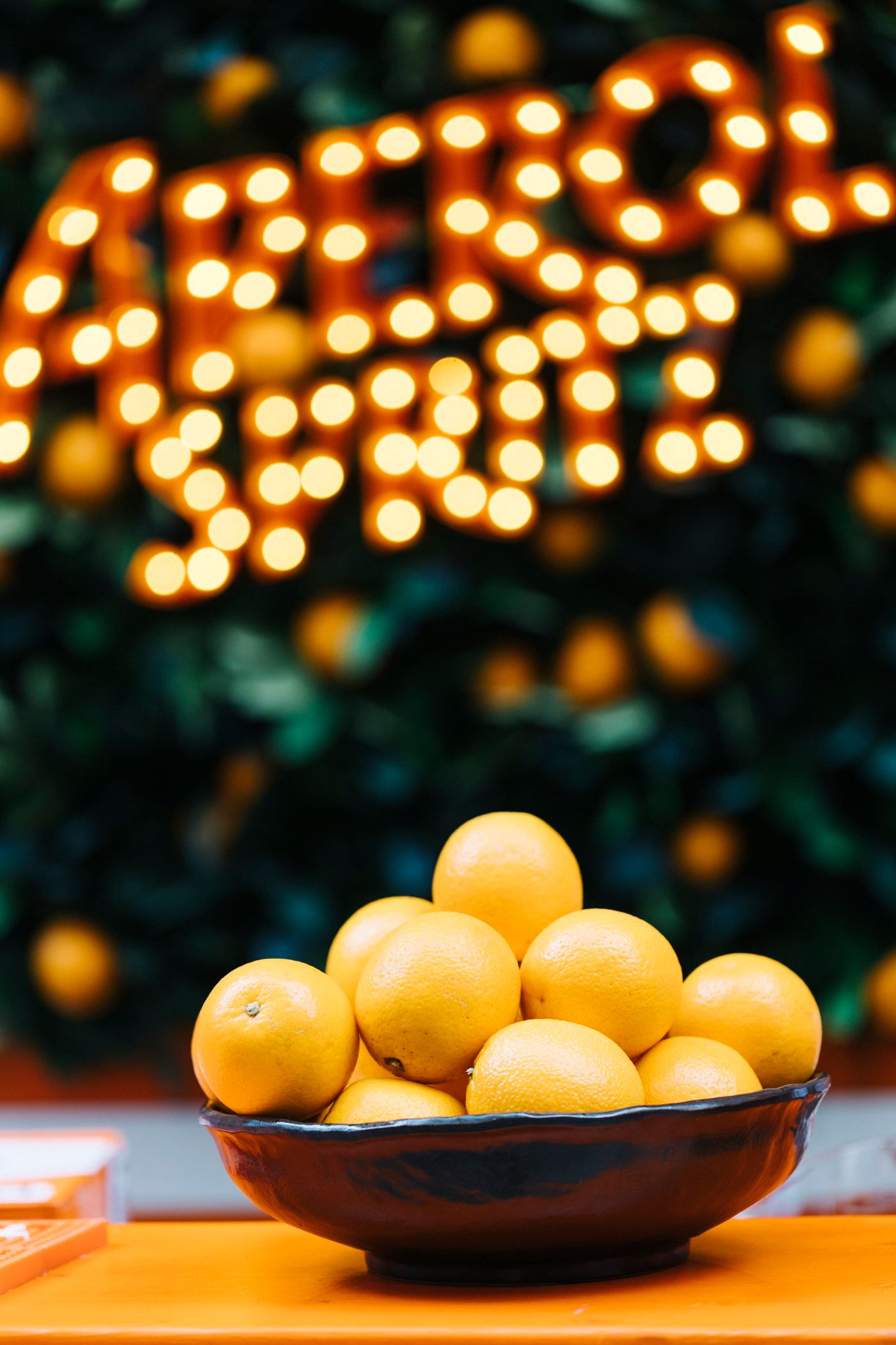 Aperol Spritz mit frischen Orangen vor beleuchtetem Aperol-Schriftzug